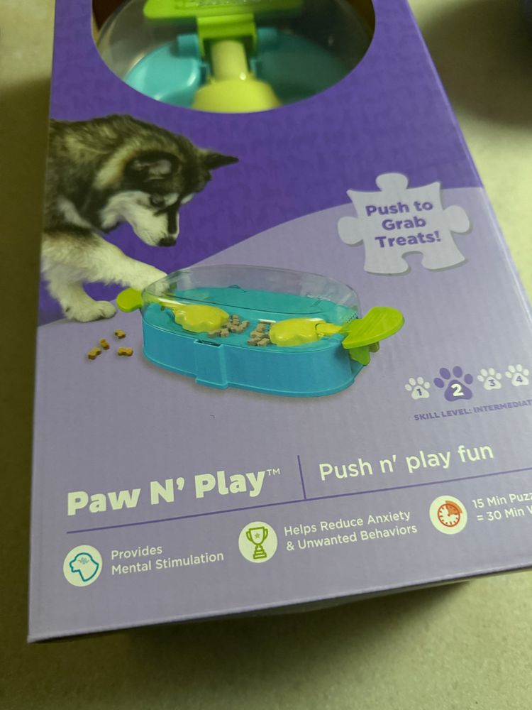 Picture of a commercial dog toy that appears to be loosely based on the mechanism involved in Hungry Hungry Hippos - a childrens game that uses levered scoops to pull marbles into a pile - but this is pulling dog treats out a hole toward the dog in question.