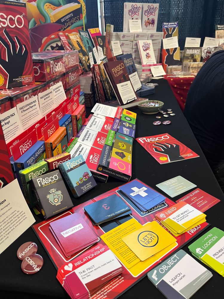 Photograph shows a convention table covered in black fabric. The table is artfully arranged with board games with Fiasco in the foreground and many of the decks available for the core RPG. In the middle of the table, there is a bowl with pinback buttons and several on the table for attendees to take. In the background, the viewer can see Star Crossed, Durance, and The Warren. 
