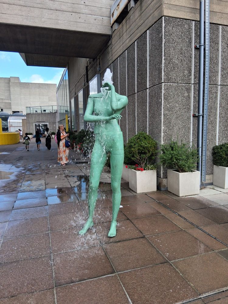 A sculpture of a figure without a head a fountain of water coming out of the neck.