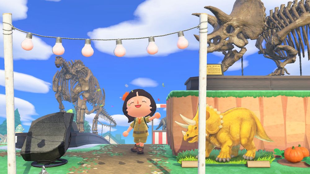 The entrance to Noodletown’s museum area featuring a string of hanging lights, an informational stone statue, a yellow triceratops standee, and a triceratops fossil behind the standee on a cliff. 