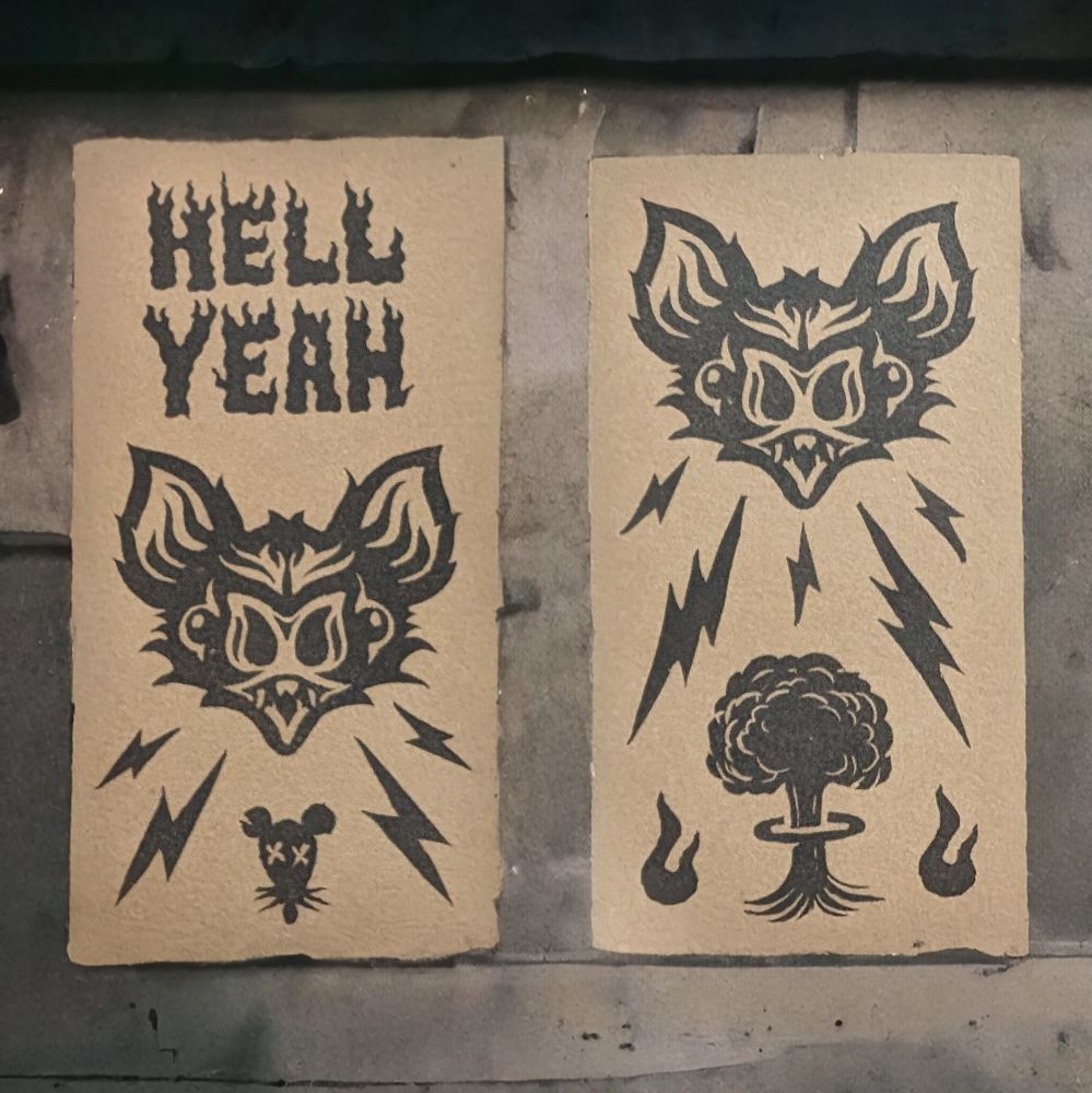 Two bookmarks sitting side by side. Both printed with h black ink in craft paper brown paper. The left on says hell yeah in flames above a bat face and the right features a bat and lightning bolts above a mushroom cloud