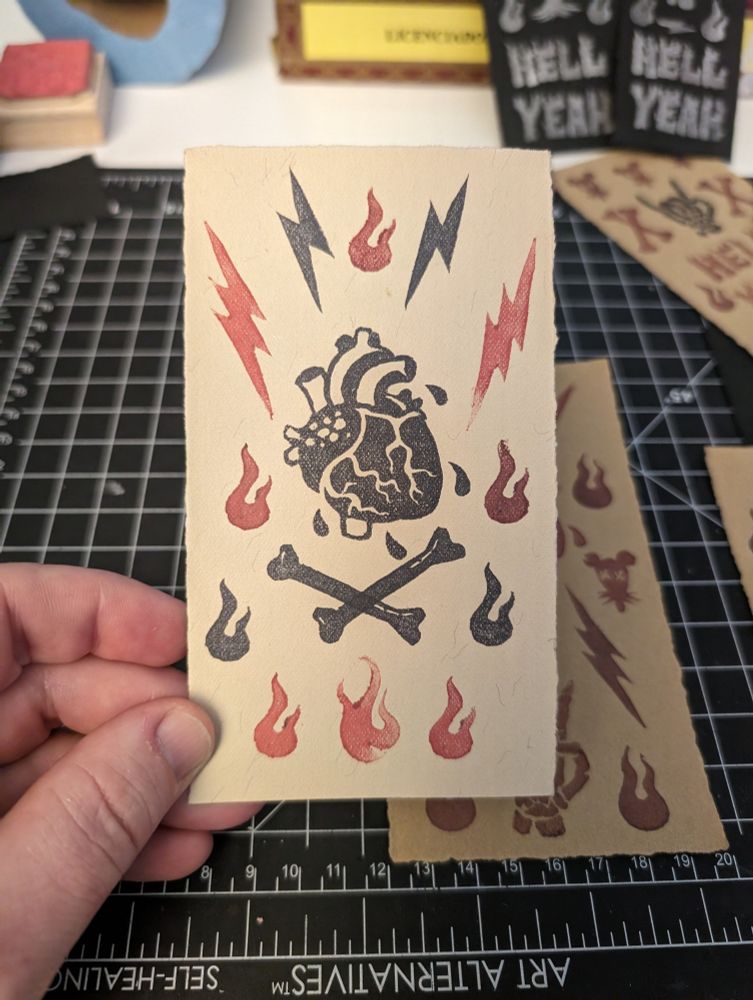 Photo of a hand holding a bookmark printed with a heart and crossbones in denim blue ink, surrounded by red lightning bolts and flames.