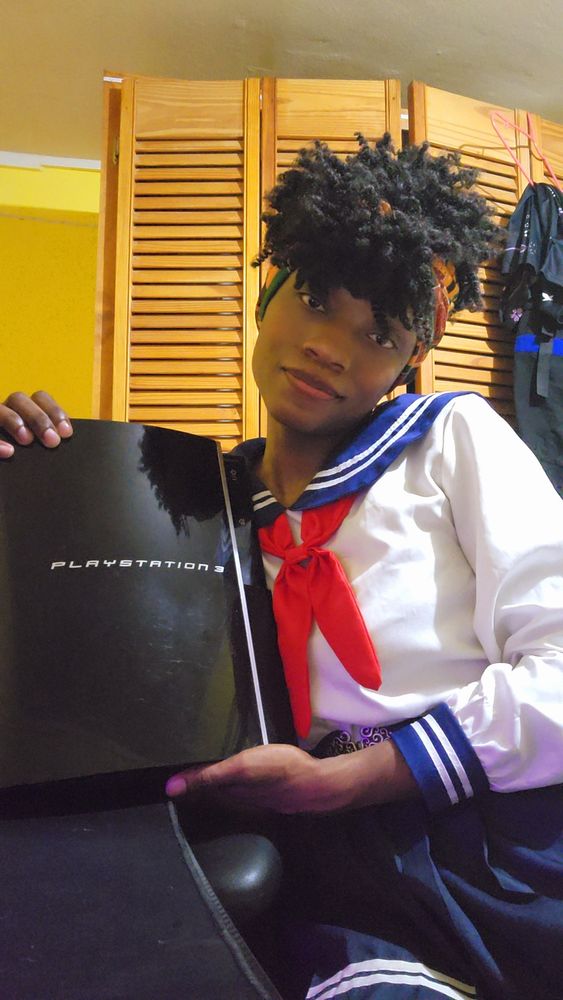A picture of a transgendered female individual wearing a white and blue sailor school uniform with a red tie and a black belt with a gold buckle around the waist whilst holding a black FAT PS3 console model CECHL01 which was jailbroken and is now non functional due to the yellow light of death. In a room with yellow walls a white tiled floor and a brown wooden closet in the background. The Playstation 3 is broken but will be held on to for future projects ;) 