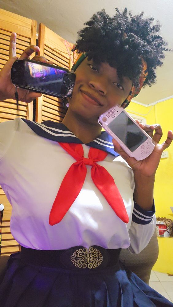 A picture of a transgendered female individual wearing a white and blue sailor school uniform with a red tie and a black belt with a gold buckle around the waist whilst holding a black, fat psp 1000 in one hand and a pink psp 2000 slim in the other whilst sitting in a Grey office chair inside of a small hot room with yellow walls a white tiled floor and a brown wooden closet in the background