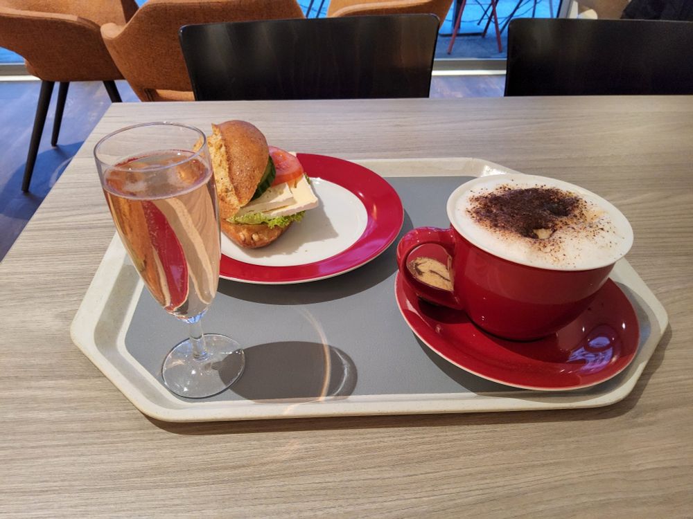 Tablett mit Tasse Cappuccino und Glas Sekt im Vordergrund und einen belegten Brötchen im Hintergrund.