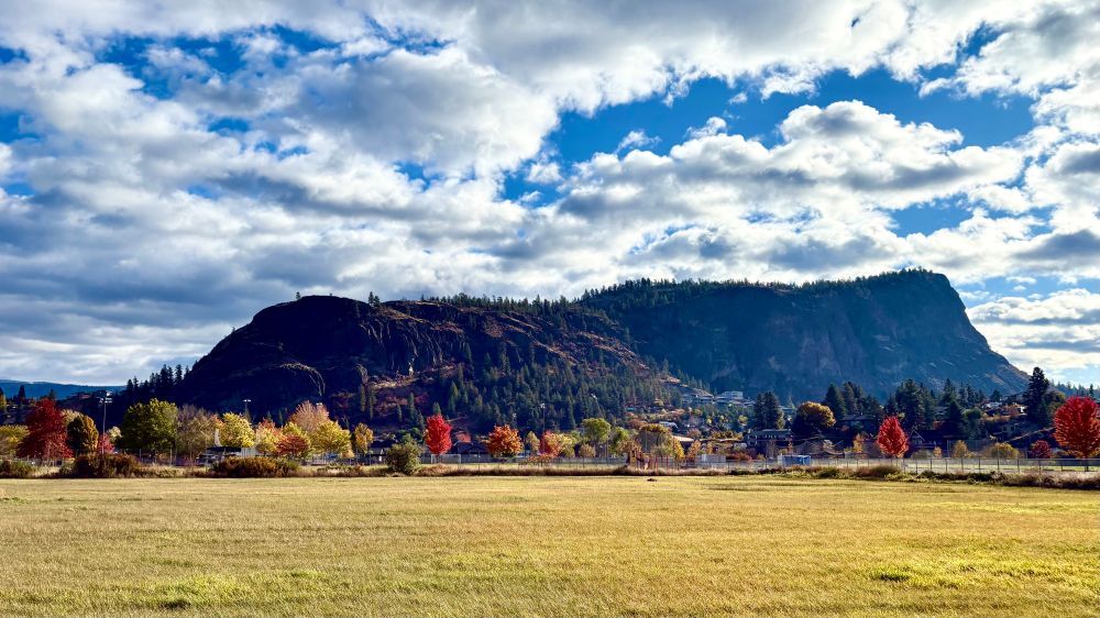 Low flat mountain against a blue sky dotted with white clouds.  Foreground is a field bordered by trees displaying fall foliage. 