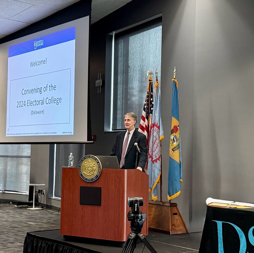 Fran O’Malley, Director of the Delaware Center for Civics Education, stands at a podium in front of flags of the United States and Delaware with a slide to his right welcoming the audience to a convening of the 2024 Electoral College. 
