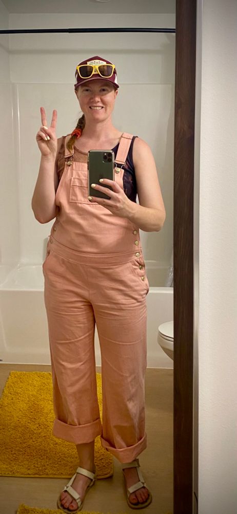 A redheaded white woman standing in a bathroom. She’s giving a peace sign and wearing a baseball cap, a pink and navy tank top, pink overalls rolled up, and white sandals. 
