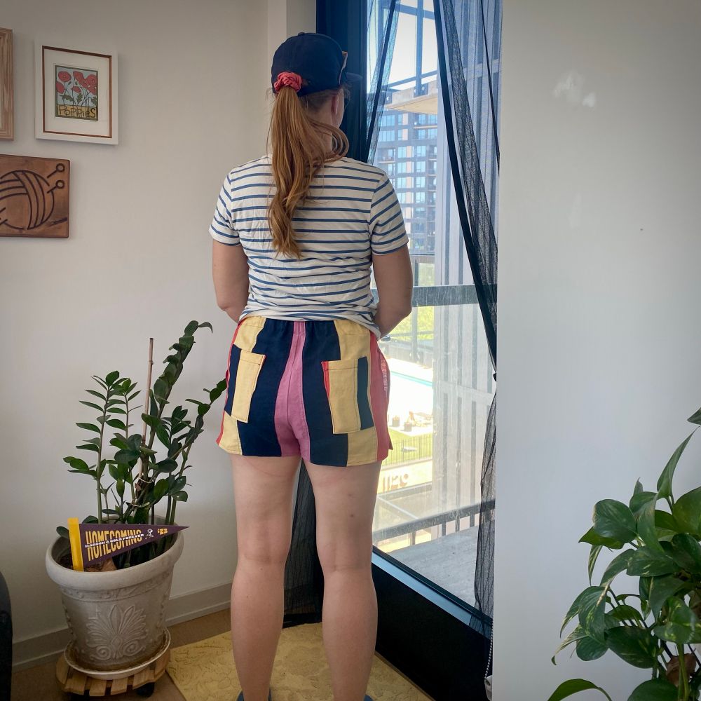 The same woman from the back. There are patch pockets on the back of the shorts, and the stripe sequence is coral, butter yellow, navy, pink. 