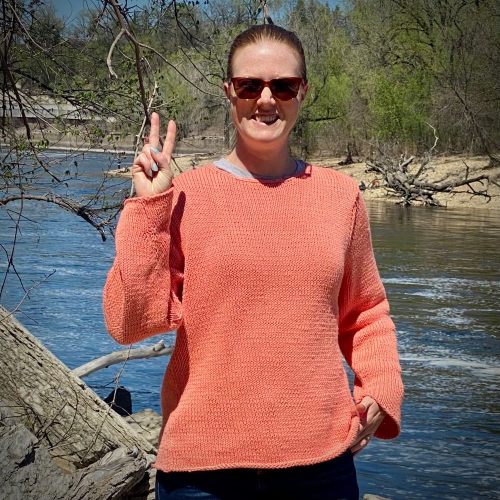 A white, redheaded woman making a peace sign with her fingers in front of a river. She’s wearing sunglasses and an oversize coral pullover.