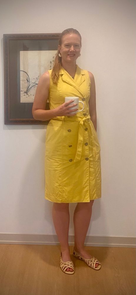 A redheaded white woman with a ponytail. She’s holding a coffee cup and standing in front of a painting. She’s wearing a yellow, sleeveless trench dress with a matching tie belt and silver button. 