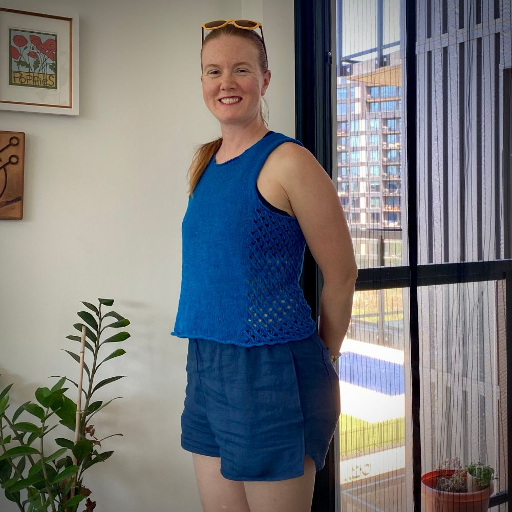A white, redheaded woman standing in front of a plant and a patio door. She’s wearing a ponytail, sunglasses on top of her head, a bright blue tank top with lacy sides, and navy elastic-waist shorts. 