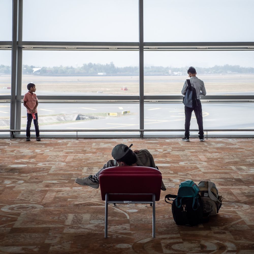 Inside New Delhi Airport: A quiet moment captured. A child stands by the large window looking out at the runway, while a man with a backpack gazes outside, lost in thought. In the foreground, another traveler lounges on a red chair, resting beside their bags on the patterned carpet. The hazy sky and empty runway stretch beyond the glass wall, giving a sense of stillness and waiting.