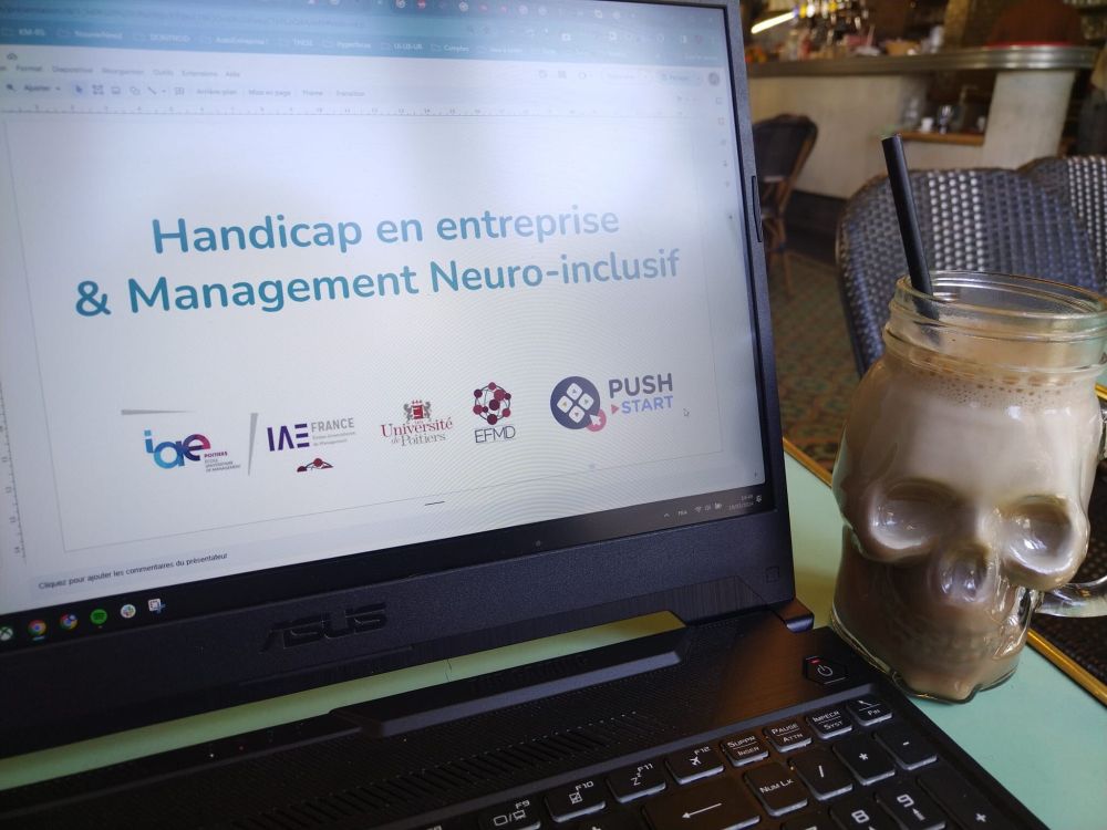 This is a photography taken in a café, of a computer with a side of coffee in a skull-shaped glass and a straw, displaying the following slide: “handicap en entreprise et Management Neuroinclusif” at a management University in Poitiers, France.