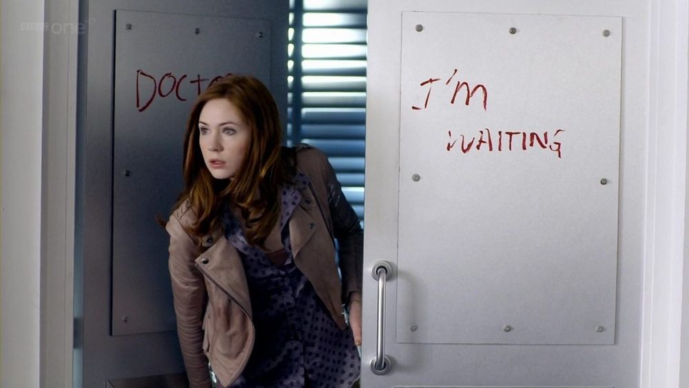 Amy Pond looks out of a door, one of two doors. One door says Doctor, and is obscured by her. The other, in fading writing, says I'm Waiting.