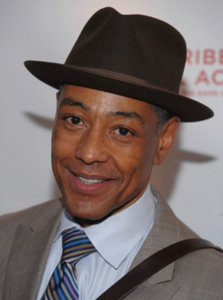 A press photo of the actor Giancarlo Esposito, shoulders up, smiling in a suit, tie and hat. 