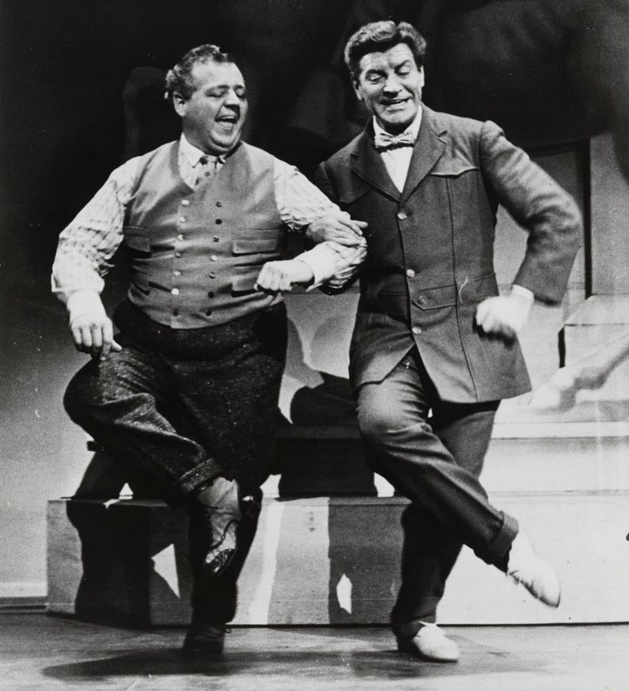 Iggie Wolfington and Robert Preston dance as "Marcellus Washburn" and "Professor Harold Hill" in The Music Man. Their arms are locked together and their right legs are raised.