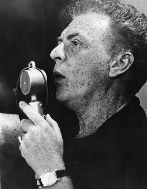 Morton DaCosta directs actors from the audience, speaking into a large microphone. His freckles are prominent on his face and neck.