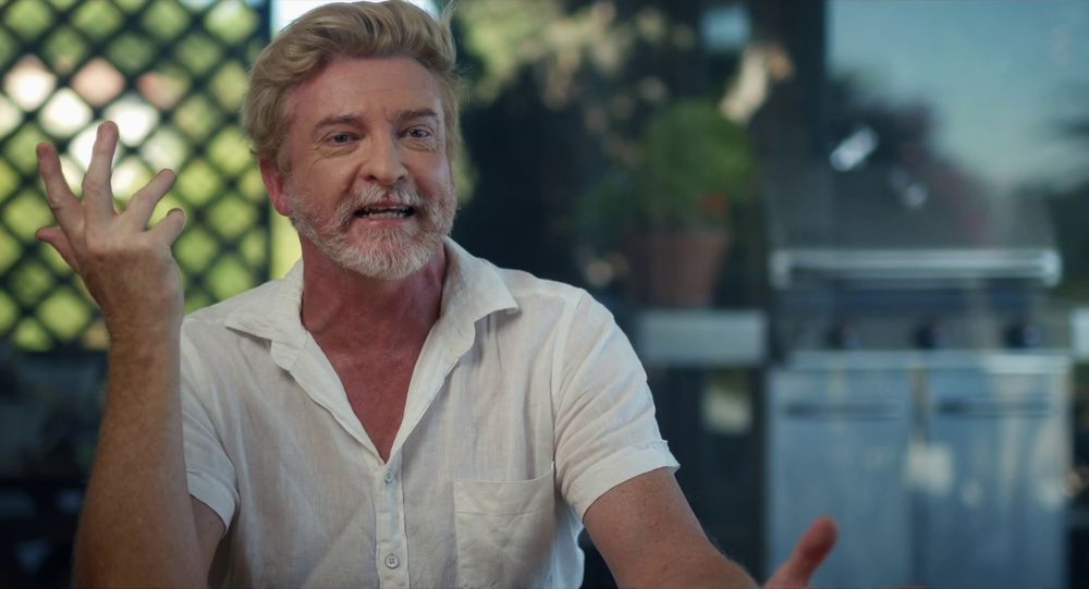 Rhys Darby in the trailer for the new documentary "But Also John Clarke". He is wearing a white linen shirt and talking with his hands.
