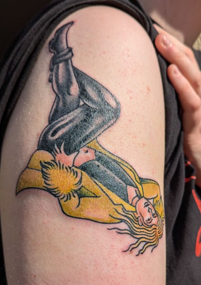 Tattoo of Lestat de Lioncourt posed like a pinup girl. He is laying on his back, one hand on his hip, the other behind his head. Legs are up at a 90° angle, crossed. He is wearing a chartreuse coat with feathered sleeves, and tight black pants.