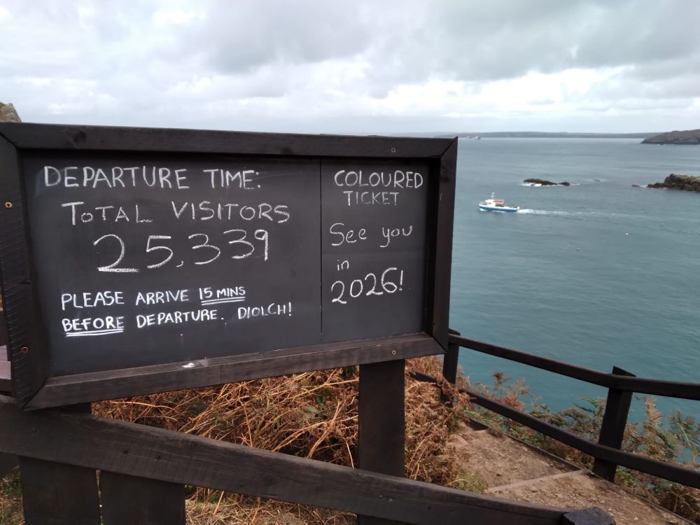 A photo of the departure blackboard which normally displays departure times. It reads "Total Visitors: 25,339. See you in 2026!" The boat can be seen leaving the bay behind.