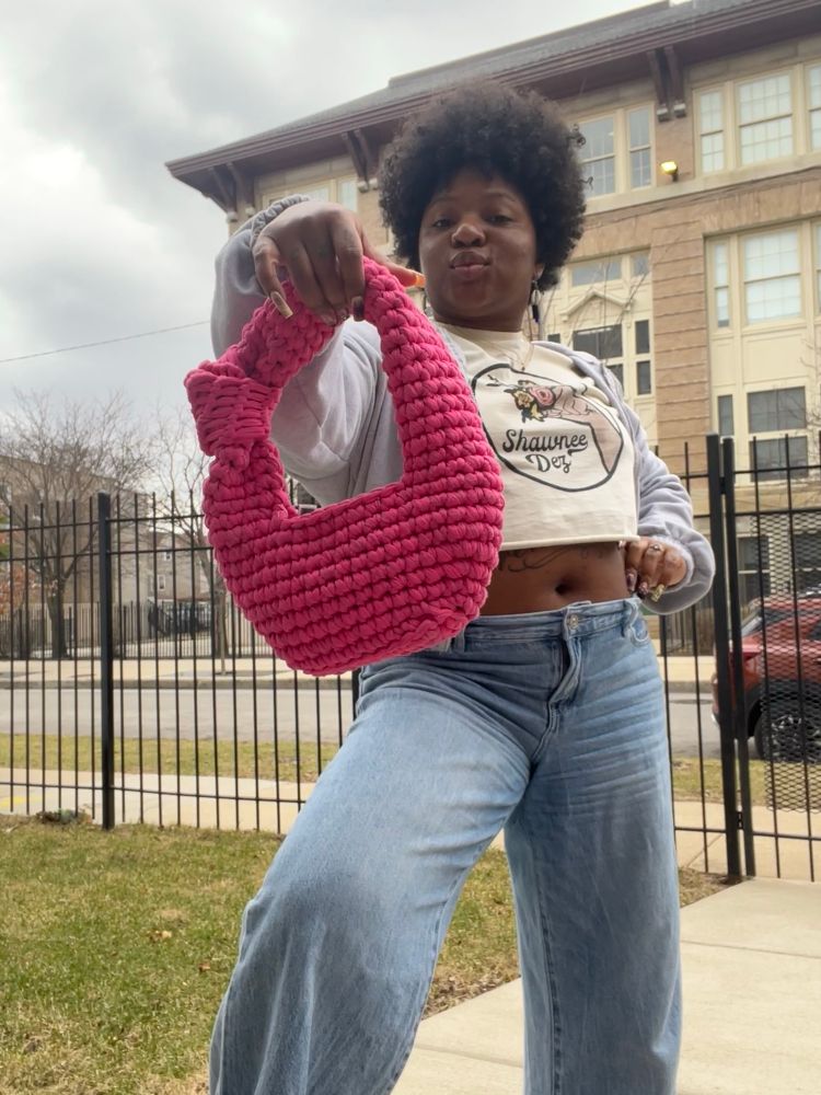 Crislin in the same setting and clothes standing closer to the camera and holding the pink crochet bag up with her other hand on her hip. She smirks proudly into the camera.