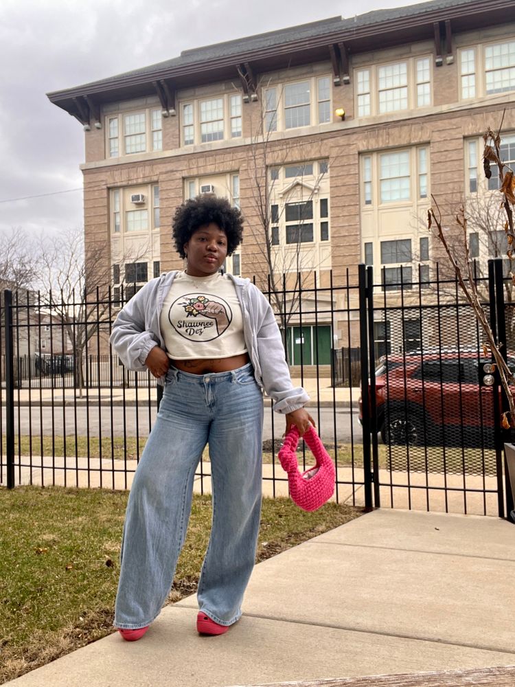 Crislin with a short curly black fro is standing in a front yard in a cropped T-shirt, heather gray cropped zip up hoodie that’s open, loose light blue jeans and pink platform crocs and holds a hot pink crochet bag in her hand