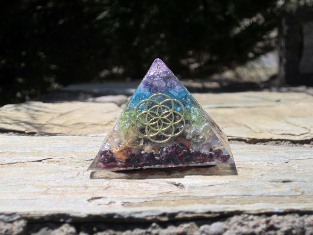 A magical-looking pyramidal gem tetrahedron beside a patio grill. The object is stacked in discrete layers with garnet, citrine, carnelian, quartz, peridot, blue topaz, and amethyst from base to apex and includes a copper wire flower-of-life motif on the front face