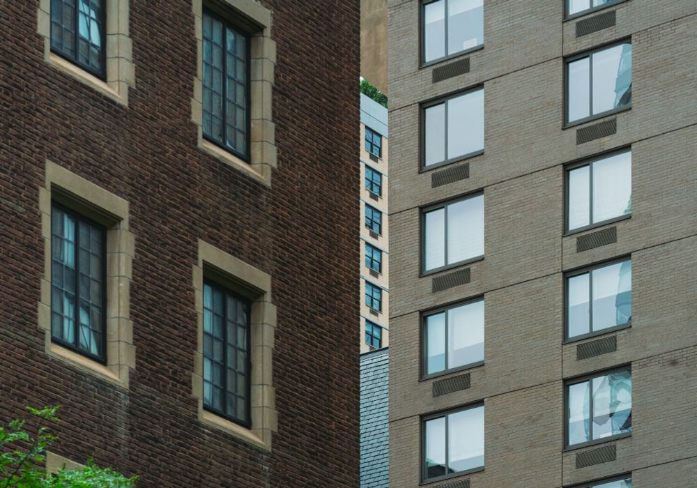 This photograph shows two buildings that neighbor each other on 59th street in the Manhattan borough of New York City. The two buildings have quite a bit of space between them, as they aren't immediate neighbors. Even though there is plenty of space around the buildings, the photograph compresses the depth into a 2-Dimensional plane. The photograph shows the south side of a brown brick building, with a cluster of dark antique pane windows that have stone borders, on the left side of the photo; and the west-facing side (that looks at the brown building) of a tan brick apartment building, with clusters of large rectangular windows reflecting light and air conditioning vents under them, to the right side of the photo. While walking a certain direction and slowly arriving to a specific position on the sidewalk, you create a frame between the buildings that shifts as you move in any direction. The tan building at the upper portion has what looks like an angled extrusion and it makes that frame have a triangular crop at the top part of the "slice" between buildings. Inside the slice is another apartment building with orange-ish brick and a single column cluster of windows. At the top of the building are green tree leaves visible, and the tan building's triangle crop extrusion makes the tree look like a tree "line". At the bottom half of the slice there is a tiled roof of a church visible, and the shape of it mimics the angled extrusion of the tan building. At the bottom left of the photo is the top portion of a tree with green leaves, which frames the visual connection made in the photograph (the two trees only see each other from this frame) as well as how the photograph was made (slowly rocking around while seemingly looking up at nothing on a city street next to a hospital 😂) 