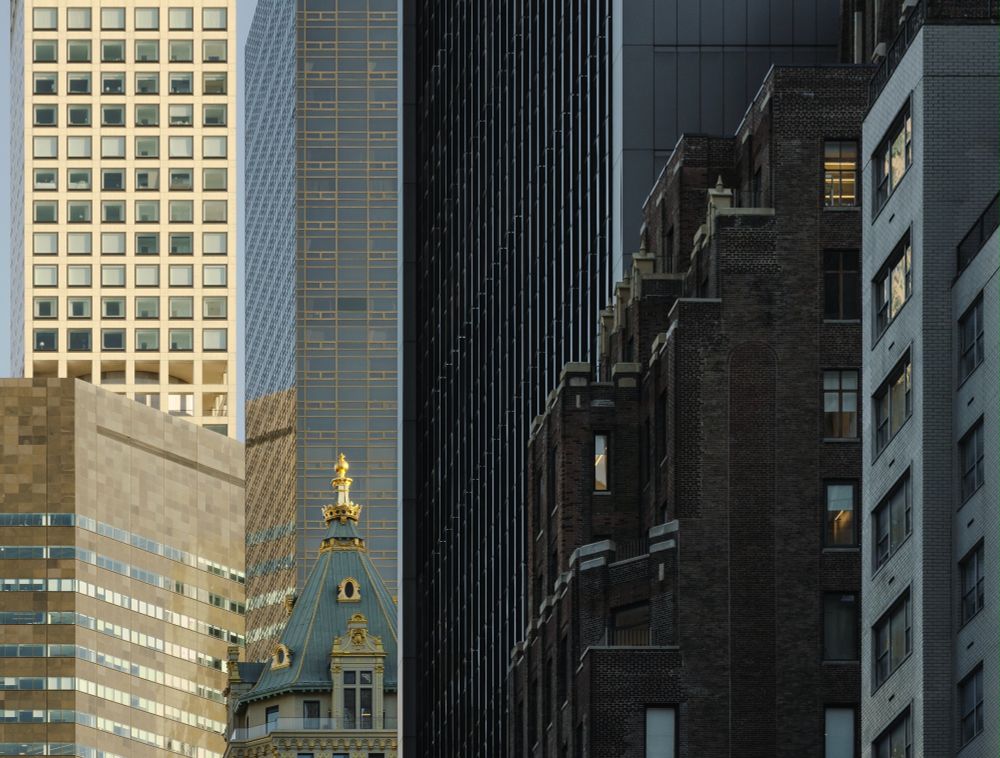 This photograph presents a series of buildings on 57th street in New York City, also known as "Billionaire's Row." Seven buildings are shown in a compressed view that visually flattens the deep landscape into a more 2 dimensional looking image. Overall, the right side of the image appears darker as the buildings are in shade while the buildings on the left side are in sunlight. Going from right to left (foreground to background), there's a sliver of of an older brick building that has supposed luxury apartments, and behind it to the left is a brown brick commercial office building owned by an equity company, and behind that is the Lefrak building which is a major landlord/developer behemoth, and then behind that is just the top of the gilded hotel  "Aman NY" which is owned by a Russian oligarch, and behind/above that is Trump tower. At the very left of the frame there are two buildings being hit with the most light, the first being the IBM building, and behind it is at the top left is 432 Park avenue, one of the most expensive places to live in NYC with many of the properties owned by mega rich foreign nationals that don't even live there, as well as other super wealthy celebrities and Industry leaders. The title "Worship the hierarchy of Capital" gives a visual metaphor of how so many parts of NYC living are inaccessible to working class and poor people, and how the rich and powerful band together and have solidarity with each other while harming the general public's ability to sustain a living. Some of them pretend to be critical of each other and some excuse warranted areas of criticism all in the goal of propping up and fighting for the system that benefits them.