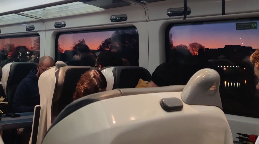 Picture taken from inside a train carriage, looking out of windows on the opposite side of the carriage to see a peach and purple sky and dark silhouettes of trees in the foreground.