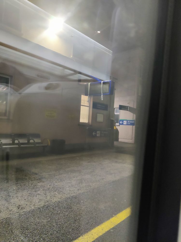 An image of a train station platform taken from inside a waiting train. It is dark outside.