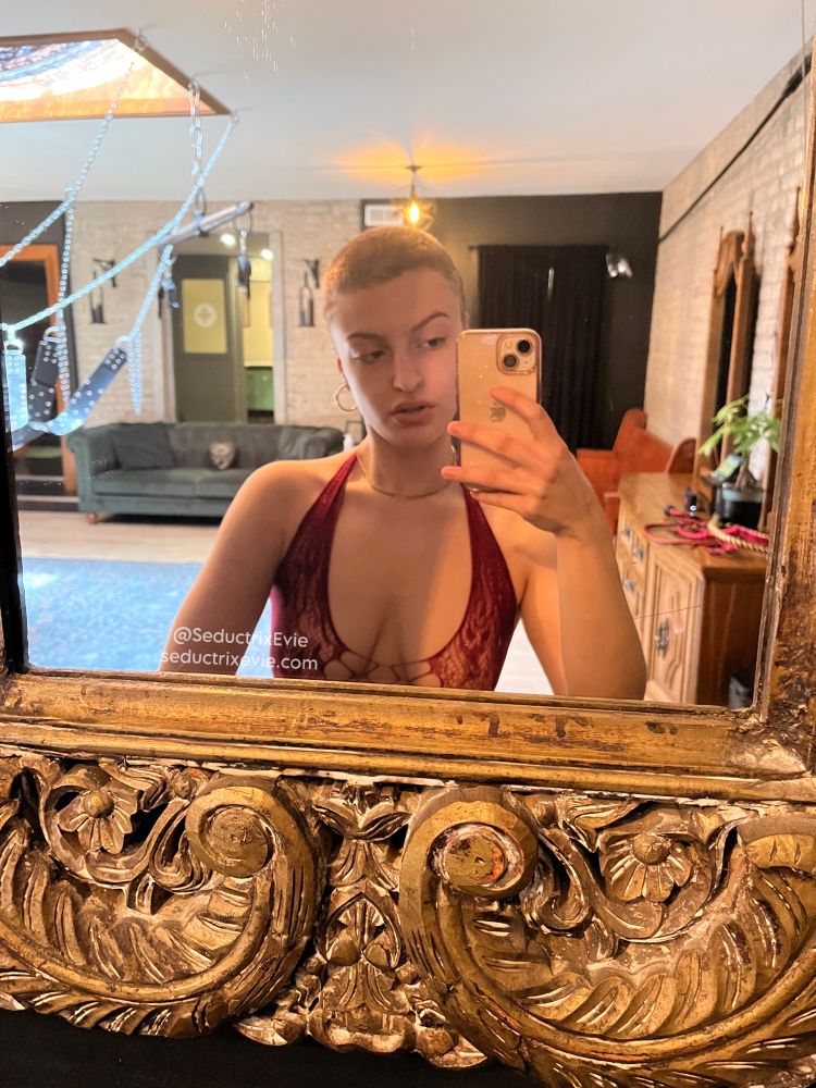 A Toronto Dominatrix taking a selfie in the dungeon mirror. Seductrix Evie has a shaved head and is wearing a red, lace bodysuit. 