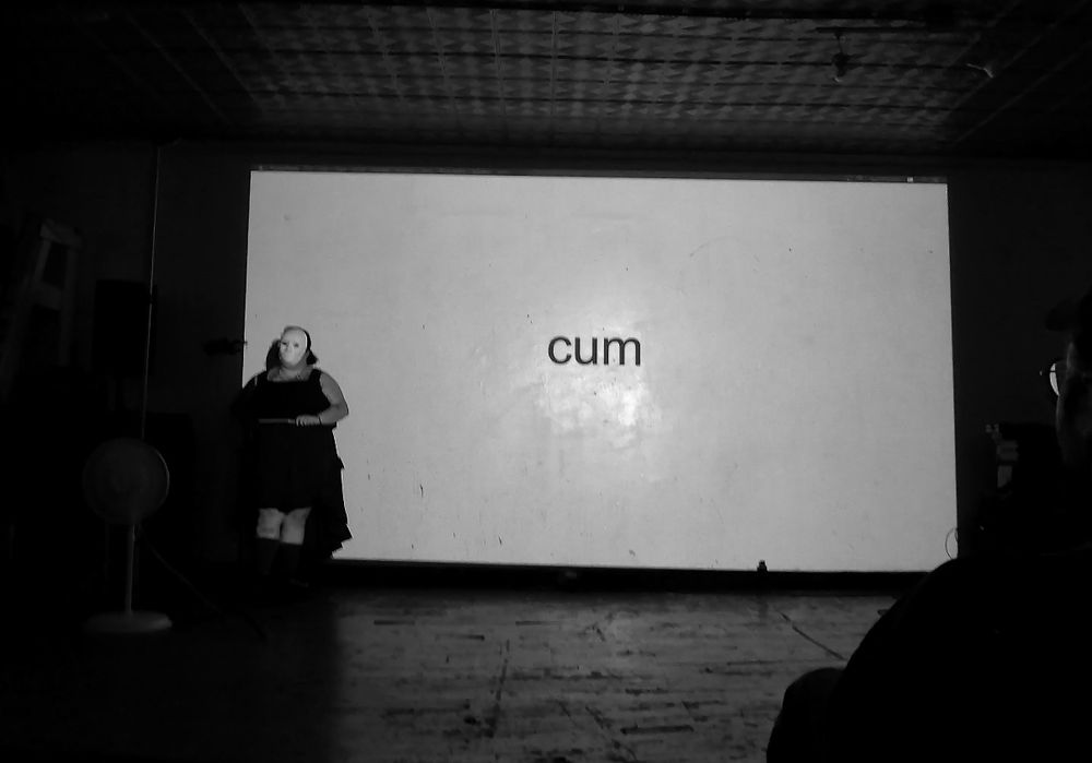 a black and white image of a figure wearing all black and a featureless white mast standing in front of a simple powerpoint of black text on white background that reads: cum
the figure is facing the audience