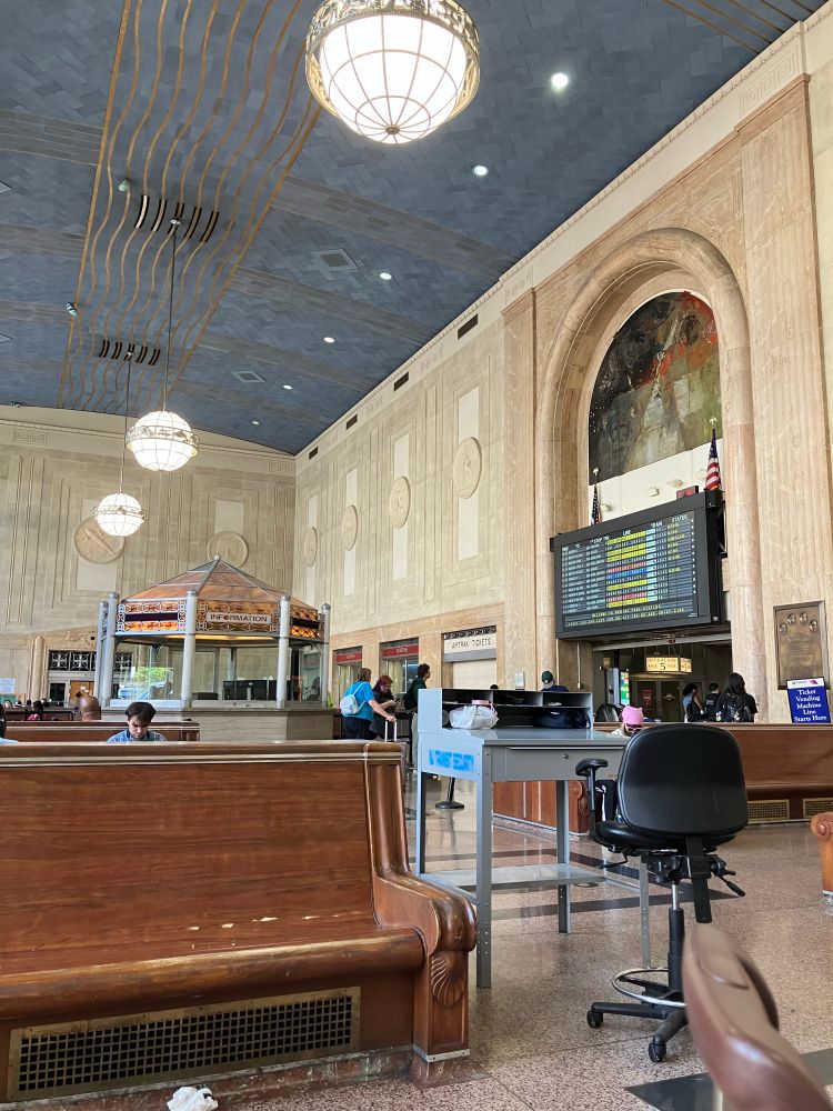 Interior of Newark Penn Station showing a wooden bench a digital board showing times a very high arched ceiling with hanging large white globe lights 