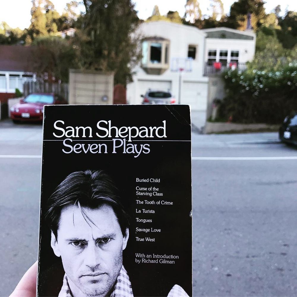 My hand holding up a copy of Sam Shelard’s book Seven Plays, which features a black and white photo of him in his 40s looking pensive, maybe even angry; in the background is the home he used to live in. The garage is in the bottom floor and the living space is on top of it.