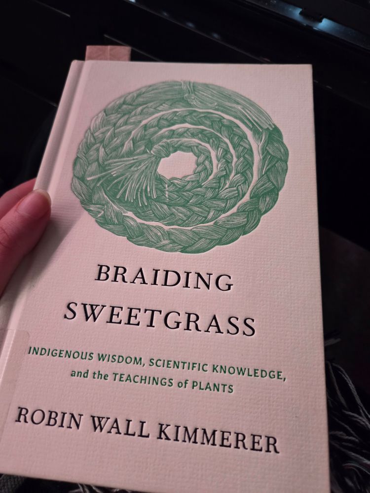 A picture of a hand holding a hardcover copy of "Braiding Sweetgrass: Indigenous Wisdom, Scientific Knowledge, and the Teachings of Plants" by Robin Wall Kimmerer. The cover features a green wood cut of braided sweet grass wound into a loose coil.