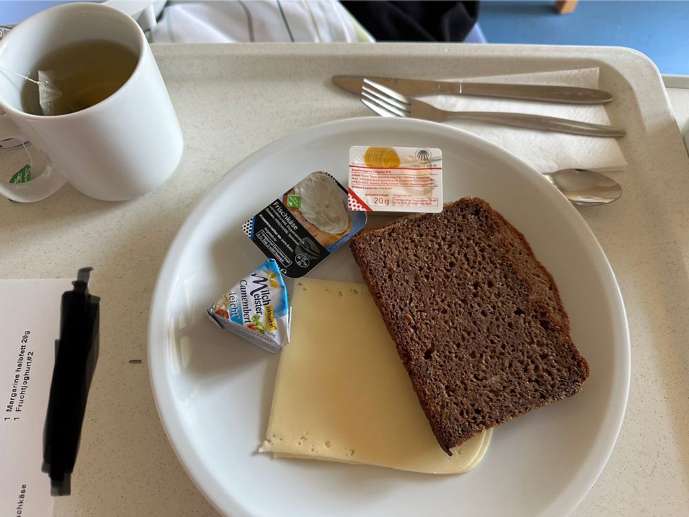 Auf dem Tablett thront ein luxuriöses Krankenhaus-Dinner: eine einsame Scheibe dunkles Brot, so kompakt, dass sie als Baustoff durchgehen könnte. Daneben eine dünne Scheibe Käse – mehr Deko als Geschmacksträger. Drei kleine Plastikportionen (Margarine, Frischkäse, Camembert) sorgen für kulinarische Vielfalt im Probierlöffel-Format. Dazu ein Becher heißes Wasser mit Teebeutel – das Highlight des Abends, wenn man Kräuteraroma mag. Besteck liegt bereit, als ob hier gleich ein Festmahl stattfinden würde. Insgesamt ein Menü, das nicht satt, sondern eher demütig macht – typisch Krankenhausküche: funktional, aber weit entfernt von lecker.