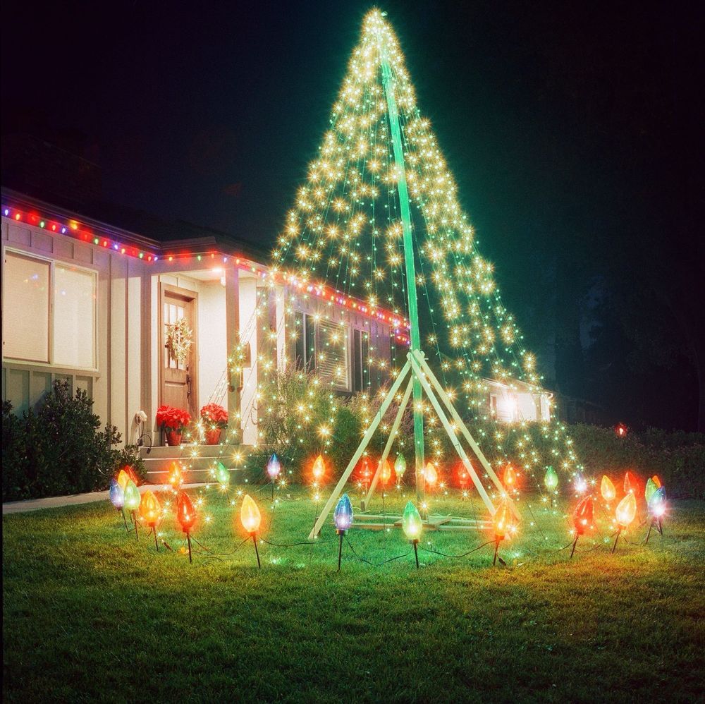 Night view of a tall Christmas tree made of green string lights, surrounded at the base with larger-than-life multi-colored Christmas bulbs. Eaves of house behind it are also lined with multi-colored lights.