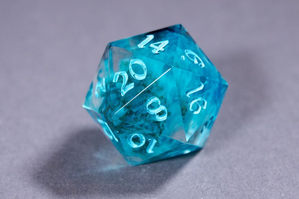 A light blue d20 with green icelandic moss and clouds of white inside, resting on a gray surface. The numbers are inked in an even lighter blue