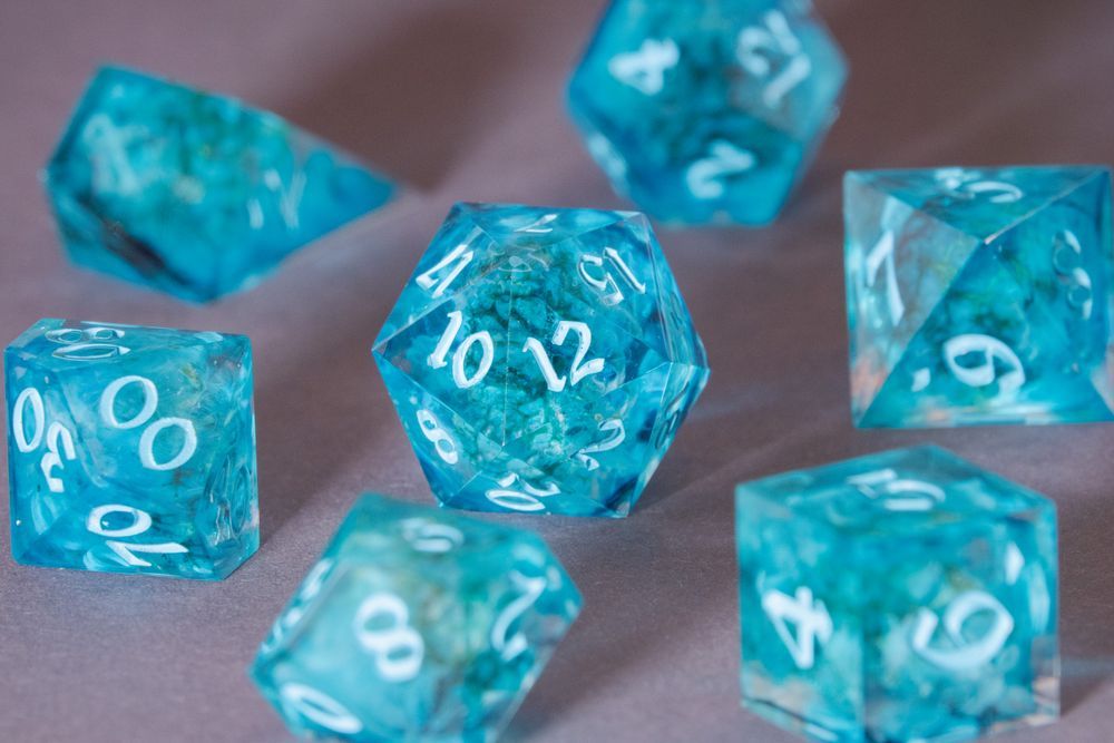 A set of light blue dice with green icelandic moss and clouds of white inside, resting on a gray surface. The numbers are inked in an even lighter blue