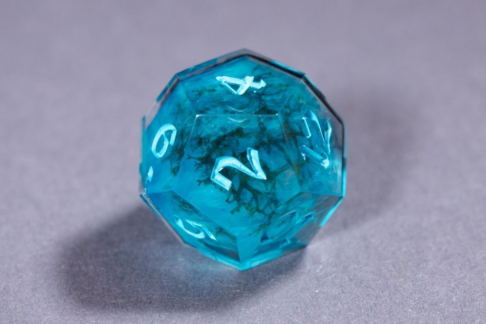 A light blue d12 with green icelandic moss and clouds of white inside, resting on a gray surface. The numbers are inked in an even lighter blue