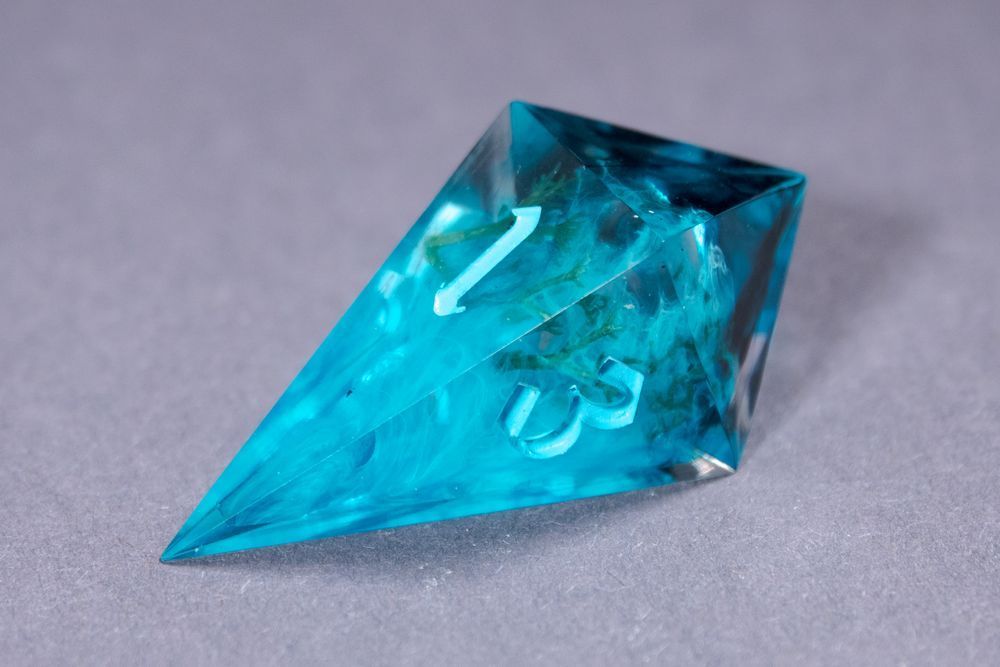 A light blue d4 with green icelandic moss and clouds of white inside, resting on a gray surface. The numbers are inked in an even lighter blue