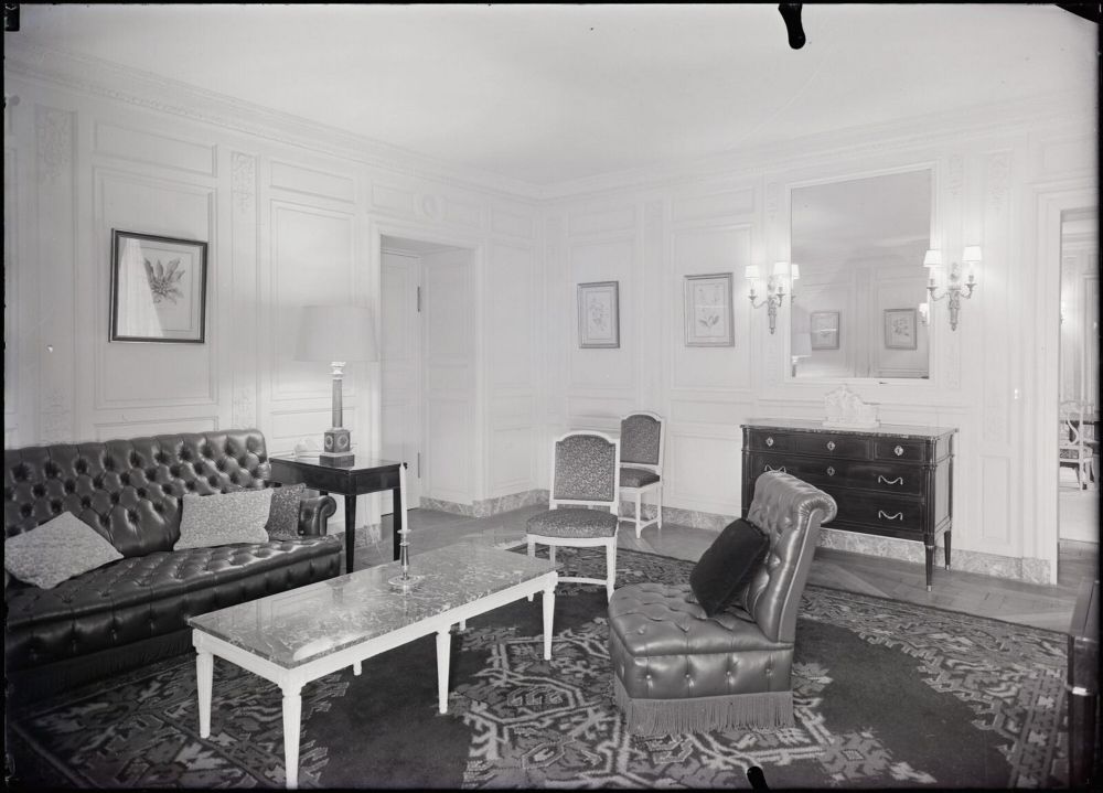 Il s’agit d’une photographie en noir et blanc montrant le salon privé du général De Gaulle, situé au premier étage de l’aile de Trianon-sous-Bois, au Grand Trianon. La pièce est élégamment décorée dans un style classique et sobre, typique de l’époque. Le mobilier est composé d’un canapé capitonné en cuir foncé, avec des coussins clairs posés dessus. Devant ce canapé se trouve une table basse rectangulaire, aux pieds blancs sculptés et au plateau en marbre. À droite du canapé, il y a un petit guéridon noir sur lequel repose une lampe à pied clair et à abat-jour blanc. À droite de l’image, un fauteuil assorti au canapé, également en cuir capitonné, est placé légèrement de biais. Il est orné d’un coussin noir. À côté, deux chaises de style Louis XVI avec dossier et assise rembourrés, en tissu à motifs, sont alignées contre le mur. Le mur en face est décoré de boiseries blanches et orné de plusieurs tableaux encadrés, probablement des dessins botaniques. Une grande commode sombre à tiroirs est appuyée contre ce mur. Au-dessus, un grand miroir rectangulaire reflète une partie de la pièce. De chaque côté du miroir, deux appliques murales avec des abat-jours blancs diffusent une lumière douce. Le sol est recouvert d’un tapis à motifs géométriques sombres et clairs, qui ajoute de la chaleur à l’ensemble. La pièce évoque une atmosphère à la fois formelle, confortable et intime, adaptée à la fonction privée qu’elle remplissait.