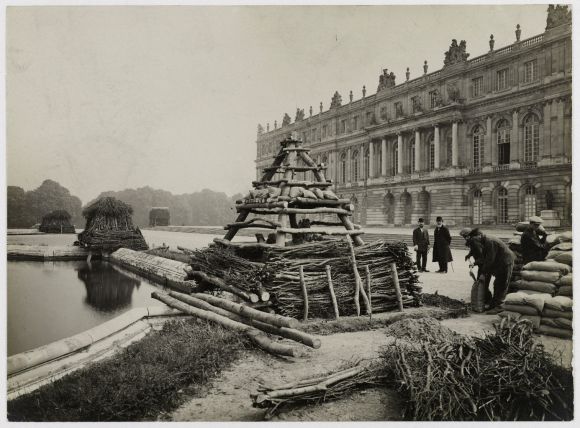 La photographie en noir et blanc montre une scène prise devant le château de Versailles durant la Première Guerre mondiale. Le bâtiment imposant du château occupe la partie droite de l’image.
Au premier plan, le long d’un bassin d’eau, on voit une structure en forme de pyramide construite avec des sacs de sable, du bois empilé et des branchages, pour protéger les sculptures. Ce type de structure est une installation de protection, typique des préparatifs militaires de l’époque. Plusieurs autres similaires sont visibles en arrière-plan autour du bassin.
À droite, quelques hommes, certains en costume civil, d'autres en habits de travail, s'affairent autour de sacs de sable. Deux hommes sont penchés, manipulant les sacs ou travaillant au sol. Un groupe d'autres personnes les observe d’un peu plus loin.
L’ambiance est grave, avec une lumière diffuse et un ciel nuageux, renforçant le caractère historique et solennel de la scène.