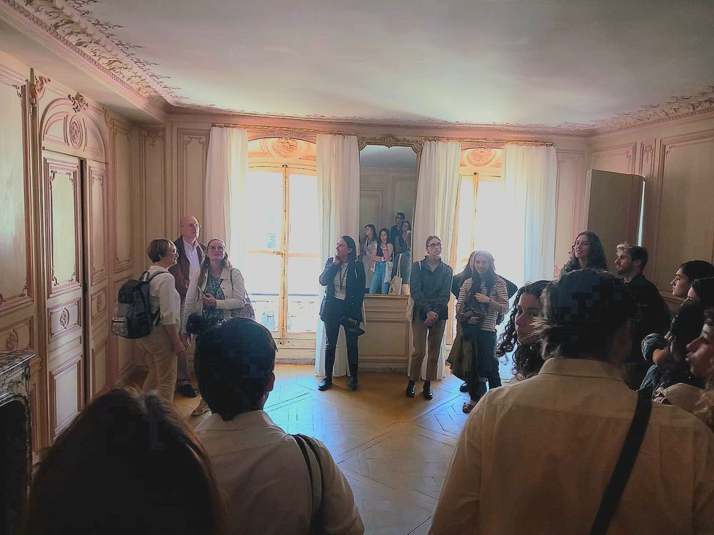 Sur cette photo prise lors d'une visite de l'appartement de Madame du Barry à Versailles, on peut voir un groupe d’une quinzaine de personnes rassemblées dans une pièce élégante à la décoration raffinée de style XVIIIe siècle. La pièce présente de hautes boiseries sculptées peintes en beige clair, avec des moulures décoratives au plafond, et un parquet au sol. Deux grandes fenêtres encadrées de rideaux blancs laissent entrer une lumière douce. Entre ces fenêtres se trouve un grand miroir qui reflète partiellement la pièce et les visiteurs. On aperçoit également une cheminée en marbre à gauche, surmontée d’un panneau sculpté. Les visiteurs, en majorité des femmes, sont debout, certains en train d’écouter, d’autres regardant les détails de la pièce. Beaucoup portent des vêtements de ville ou de tourisme, avec des sacs ou des sacs à dos. L’ambiance est calme, studieuse, comme dans une visite guidée culturelle. On sent que les gens sont curieux et intéressés par le lieu chargé d’histoire. La lumière naturelle venant des fenêtres crée un jeu d’ombres et de contre-jour, ce qui rend certaines silhouettes en partie sombres, accentuant le contraste entre la clarté extérieure et l’éclairage intérieur tamisé.