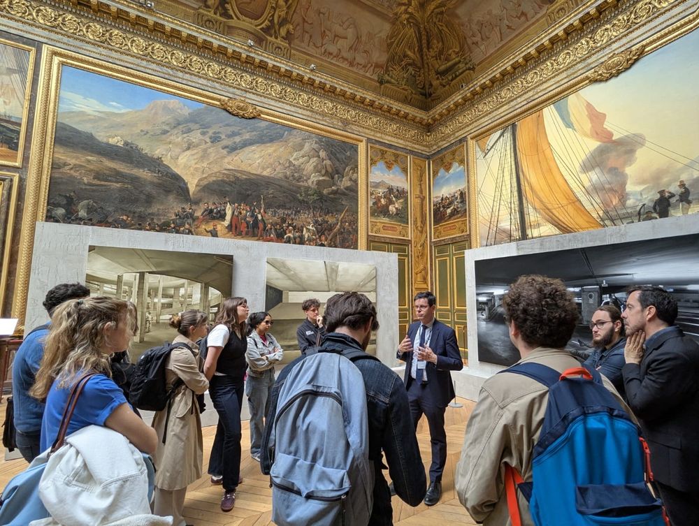 Sur cette image, on voit une scène vivante et immersive prise lors d’une visite guidée au Château de Versailles. Le président du château, élégamment vêtu d’un costume sombre avec cravate, se tient au centre du groupe et s’adresse à une douzaine de visiteurs attentifs. Il s’agit d’une visite de l’exposition de l’artiste Guillaume Bresson, dont le président est également le commissaire.
Les visiteurs, hommes et femmes, sont jeunes pour la plupart, habillés de manière décontractée, certains avec des sacs à dos, écoutant avec intérêt. Ils forment un demi-cercle autour du président, dans une salle richement décorée.
La salle elle-même est grandiose, typique du style Versailles du 19e siècle : parquet en bois, murs dorés et moulurés, plafonds peints et très hauts, avec de grandes fresques historiques représentant des batailles.
L’exposition contemporaine de Guillaume Bresson contraste subtilement avec le décor classique. Deux grandes œuvres peintes ultra réalistes sont visibles sur les murs : elles montrent des scènes modernes d’un parking souterrain, avec une ambiance urbaine et sombre. Ces œuvres, encadrées dans des structures modernes en béton clair, semblent volontairement s’opposer aux peintures classiques du 19e siècle pour créer un dialogue entre passé et présent.