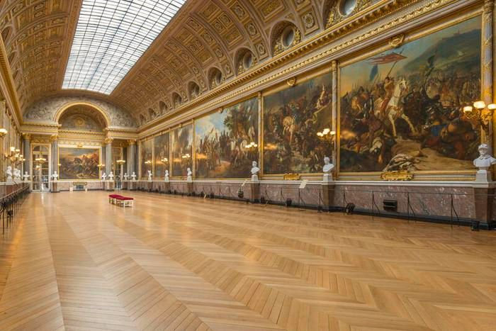 Vous êtes dans une immense galerie historique appelée la Galerie des Batailles, située dans le château de Versailles en France. La pièce est très longue, avec un plafond voûté spectaculaire et orné de moulures dorées complexes. Le plafond est en grande partie constitué d’une verrière qui laisse entrer une lumière naturelle abondante.

Sur le mur de droite, toute la longueur est couverte de gigantesques tableaux représentant des scènes de batailles célèbres de l’histoire de France. Chaque tableau est encadré de dorures massives, et les scènes sont très animées, avec des chevaux, des soldats, et beaucoup de mouvement. Devant ces tableaux, une série de bustes en marbre blanc sont alignés, représentant probablement des figures militaires ou historiques.

Le sol est recouvert d’un parquet en bois clair, disposé en chevrons, qui donne une sensation de chaleur et d’élégance. Au fond de la galerie, on distingue un grand tableau central sur un mur arrondi, toujours dans un style très dramatique et héroïque.

L’ensemble de la pièce respire la grandeur, la gloire militaire et le faste royal du 19e siècle.