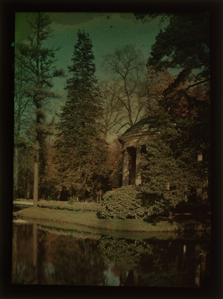 L’image montre un petit édifice élégant, appelé le Temple de l’Amour, situé dans un paysage de jardin soigneusement dessiné. Le temple est placé au centre de la composition. Il est rond, de style antique, avec douze colonnes blanches qui soutiennent un toit en forme de dôme. Il ressemble à un petit monument grec ou romain, simple mais raffiné.

Au milieu de ces colonnes, à l’intérieur du temple, on aperçoit une statue de marbre clair représentant l’Amour — un jeune garçon nu, aux ailes repliées, tenant un arc : c’est Cupidon. Il est debout sur un piédestal, au centre exact de la rotonde.

Le temple repose sur une plateforme circulaire, entourée de marches basses. Autour du bâtiment, la végétation est abondante mais ordonnée : on voit de grands arbres à feuillage dense, des buissons, et des parterres d’herbe.

Un petit cours d’eau traverse le paysage au premier plan. Un pont de pierre simple ou une passerelle en bois permet de le franchir pour accéder au temple. La scène est baignée d'une lumière douce, parfois dorée si c’est une aquarelle ou une gravure colorée, évoquant un matin ou une fin d’après-midi paisible.

À l’arrière-plan, on distingue d'autres éléments du jardin : une prairie dégagée, des arbres en bosquet, peut-être une allée. Aucune personne n’est visible ou parfois seulement de petites silhouettes au loin, ajoutant à l'impression de calme et d'isolement du lieu.

Le style général de l’image est romantique et pittoresque. Elle cherche à transmettre l’idée d’un lieu idéal, intime, harmonieux — un refuge champêtre voulu par Marie-Antoinette loin de la cour.