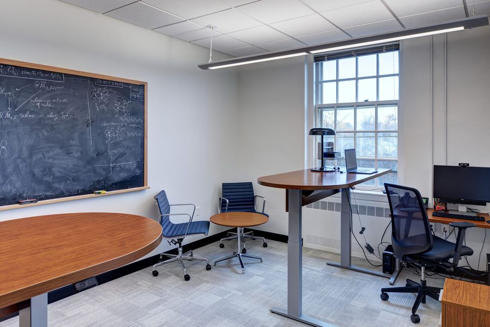 Office located at IAS. There’s a blackboard on the wall with mathematical formulas with painted white walls. There’s are multiple tables, chairs for visitors and in the corner a computer with a table setup next to a standing desk with a laptop and lamp.